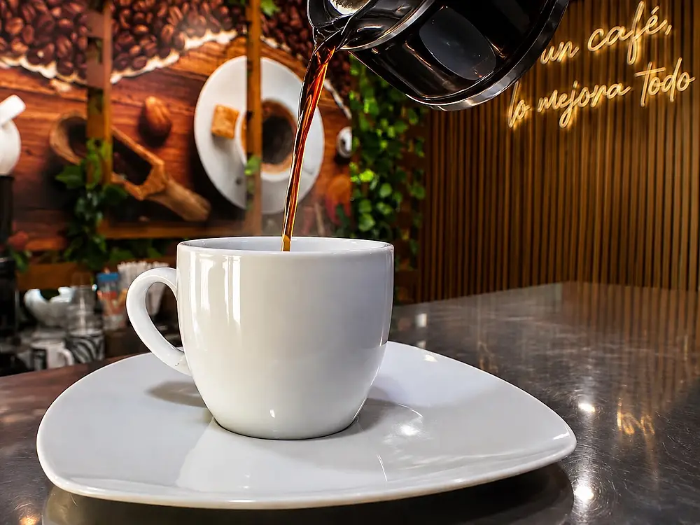 Taza de café blanca sobre la barra de RanCholaos, con el mural de granos de café y el aviso de neón «un café, lo mejora todo» al fondo